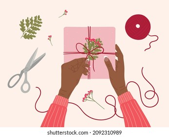 Valentine's day concept. Black female hands open gift box wrapped into kraft paper. Top view table with romantic eco-friendly packaging, hearts, scissors, flowers. Hand drawn vector illustration