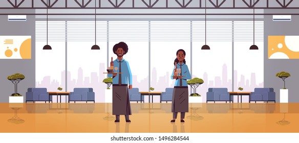 waiters couple holding bottles and glasses with champagne and wine on tray african american woman man restaurant workers in apron carrying alcohol drinks modern cafe interior full length horizontal