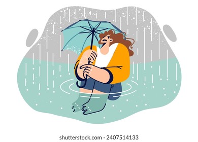 Wet woman with umbrella sits in puddle during rain and risks catching cold and needs roof over head. Frightened girl was caught in autumn rain, which caused flood and flooding of buildings.