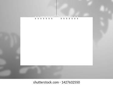 A white blank calendar or notebook on a spiral hangs on a thread. The shadow of the Monstera plant falls on the wall behind. Realistic mockup for design