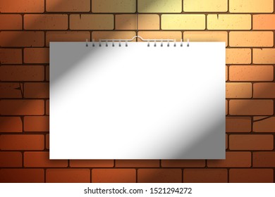 A white blank calendar or notebook on a spiral hangs near the red brick wall. Realistic mockup for design with shadow from a window