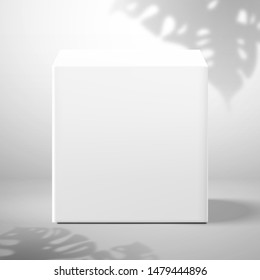 A white blank cubic box. Front view. Shadow from a Monstera plant on the wall in the background. Mockup for design