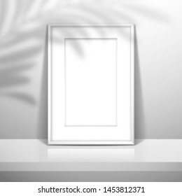 A white blank frame with passe-partout for a picture or poster stands near the wall on the shelf. The shadow of the leaves of the palm tree is on top. Mockup for design