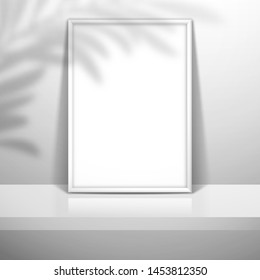 A white blank frame for a picture or poster stands near the wall on the shelf. The shadow of the leaves of the palm tree is on top. Mockup for design
