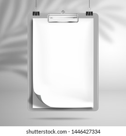 White blank notebook with a bent top sheet is clamped in the clipboard and hanging on the clips above the surface. The shadow of the palm tree leaf falls on the wall behind. Mockup for design