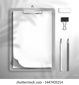 White blank notebook or sketchbook with a curved top sheet is clamped in the clipboard, two pencils, binder and an eraser. The shadow of the leaves of the palm tree is on top. Flat lay mockup