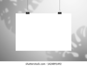 White blank poster hang on clips. The shadow of the Monstera plant falls on the wall behind.  Mockup in horizontal orientation
