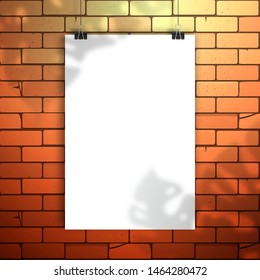 White blank poster or sheet of paper hangs on the clamps on red brick wall. The shadow of the leaves of the Monstera plant is on top. Mockup for design