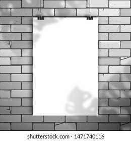 White blank poster or sheet of paper hangs on the clamps on brick wall. The shadow of the Monstera plant is on top. Mockup for design