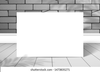 A white blank rectangle or sheet of paper is standing near a brick wall on a plank floor. The shadow of the palm tree is on top. Mockup for design