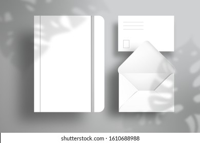 A white notebook with an elastic band, a postcard and an open envelope. Realistic branding design with shadow from the leaves of a Monstera plant. Flat layout of white objects