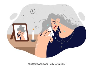 Widow woman is crying standing near portrait of deceased husband and wiping tears from face with handkerchief. Elderly lady widow in mourning clothes remembers partner who died due to fatal illness.