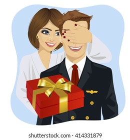 wife in white bathrobe covering her husband's eyes wearing airline pilot uniform standing behind him with gift