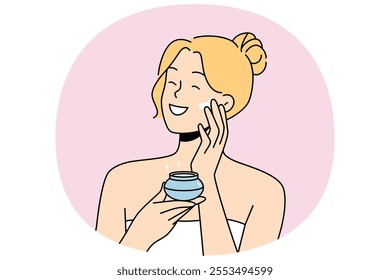 Woman applies anti-aging cream to face to stay beautiful, standing in bath towel after shower. Happy blonde girl smiles and uses cosmetic cream to help prevent wrinkles and pimples.