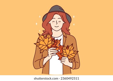 Woman with autumn leaves in hands smiles standing in stylish casual clothes and hat. Cheerful young girl rejoices at onset of autumn season and collects beautiful foliage from trees