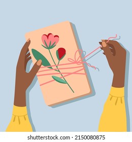 Woman with beautiful dark skin is holding a box in her hands. Gift box on the table, top view. African female hands untie the ribbon on present. The box from kraft paper is decorated with flowers