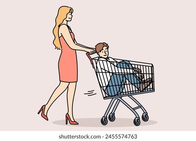 Woman carries man in supermarket carts, for concept of buying boyfriend and financially motivated marriages. Girl gives capricious boyfriend ride on buyer cart because guy doesnt want to go shopping