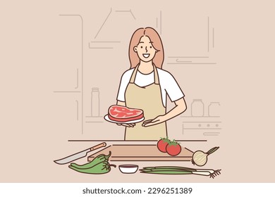 Woman cook holding raw meat steak standing near kitchen table with cutting board and vegetables. Girl offers to cook beef meat and looks at camera calling for recipe for delicious dish