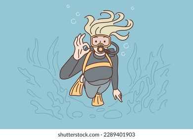 Woman diver swims underwater in scuba gear and goggles showing OK gesture exploring underwater world. Girl is diving using equipment for professional divers and following bottom of ocean with algae