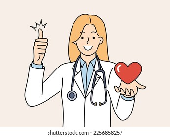 Woman doctor with heart in hand smiling and showing thumb up rejoicing at good health of patient. Girl doctor with stethoscope around neck works in cardiology department clinic