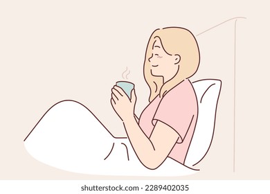 Woman is drinking coffee sitting in bed under blanket enjoying morning weekend and good mood. Girl with mug of coffee has breakfast in bedroom, drinks invigorating drink to wake up faster and smiles