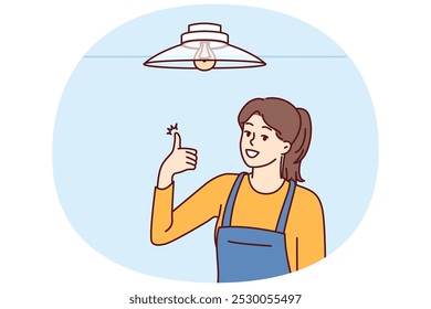 Woman electrician showing thumbs up after fixing chandelier or replacing light bulb under ceiling. Girl in overalls works as electrician efficiently performing tasks of setting up electricity