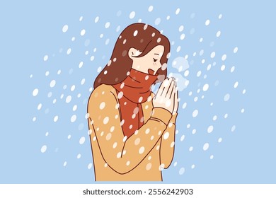 Woman enjoys winter weather, stands under falling snow flakes in sweater and scarf. Positive girl rejoices at onset of winter and approach of christmas holidays, blowing on hands to keep warm