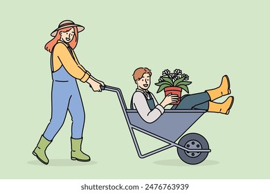 Woman farmer pushes man inside cart, enjoying good atmosphere in agro company team. Flowers in hands of farmer for planting seeds in fields and floriculture and getting large harvest