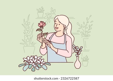 Woman florist works in flower shop composing beautiful festive bouquets for customers. Happy girl in apron makes career as seller or florist standing behind counter and holding rose in hand