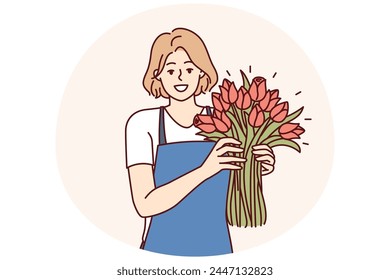 Woman flower seller stands with luxurious bouquet and smiling looks at camera enjoying work of florist in flower shop. Positive girl in apron holds fragrant bouquet with scarlet buds