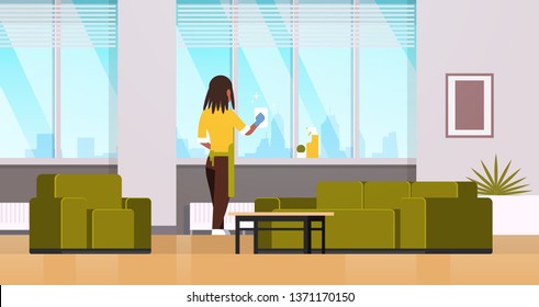 woman in gloves and apron cleaning windows with rag cleaner spray rear view african american housewife doing housework concept modern living room interior horizontal flat full length