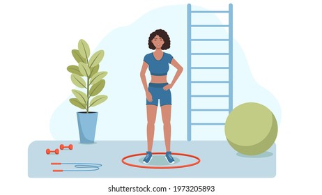 A woman in good shape is standing in the gym, next to a hoop , dumbbells, a jump rope and a fitball , in the background a ladder .
