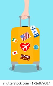 Woman hand is holding Yellow Travel bag with sticker and banner on blue background. Suitcase with airline tag. Around the world, Road trip vacation. Tourism, Journey concept. 