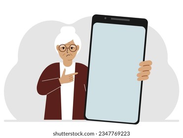 A woman holds a mock-up of a large smartphone with a blank screen and with his second hand points to the screen of the phone.