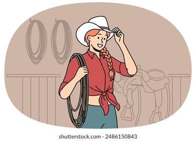 Woman jockey stands in stable wearing cowboy hat and holding long rope waiting for horses to appear. Girl rancher dressed in cowgirl style smiling when you cheer for jockey at races