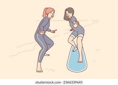 Woman learns surfer along with instructor, standing on sandy beach on surfboard, and practicing to take right posture. Girl wants learn how ride surfboard during summer trip to ocean