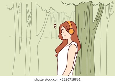 Woman listens to music through headphones standing in park near tall tree and enjoying good weather and fresh air. Teenager girl listens to music relaxing alone in nature after hard day at school