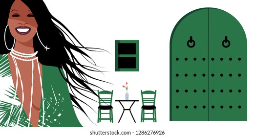 Woman with long brown hair and large earrings, on background of typical Spanish Mediterranean style village. Green door and window, chairs and small table with vase on white walls.
