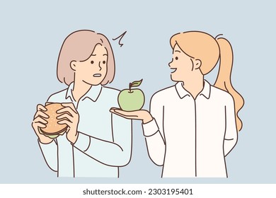 Woman nutritionist offers apple to girl who eats fast food with high content of cholesterol and fat. Nutritionist explains to patient about benefits of fruits and timing of burgers and sandwiches