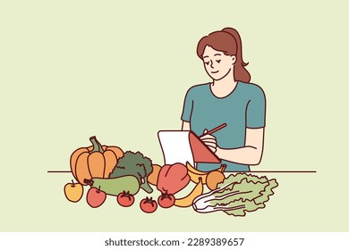 Woman nutritionist is standing near table with vegetables and making notes in notebook, making plan for new diet to detoxify body. Girl nutrition specialist uses checklist to mark diet ingredients 