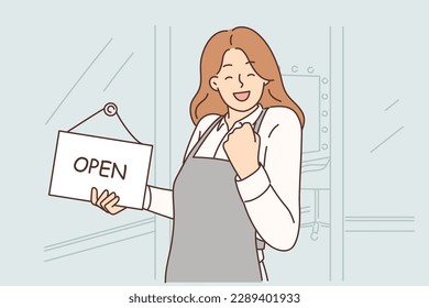 Woman owner of beauty salon makes victory gesture standing near front door with sign open on day of business launch. Girl stylist opens own business wanting to become successful entrepreneur