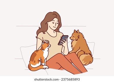 Woman owner of dog and cat sits on couch and plays on phone, enjoying spending time with pets. Girl strokes pets in need of affection and attention, to advertise shelter of domestic animals