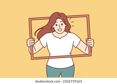 Woman peeks out of picture frame wanting to take creative shot for photo album. Positive girl in casual clothes with smile poses for photo shoot to get portrait for blog or social networks