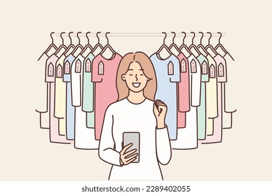 Woman with phone near hanger with T-shirts rejoices in sale and opportunity to buy clothes at discounts. Girl uses smartphone for bargain shopping through online application with discounts