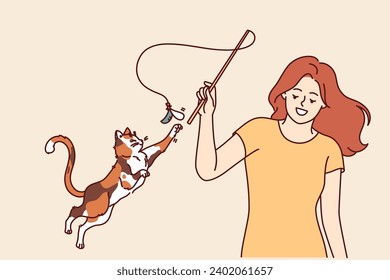 Woman plays with cat and waves bow on string, feeling pleasure thanks to presence of pet. Playful cat jumps up and stretches out paw wanting to catch kitten toy bought at pet store