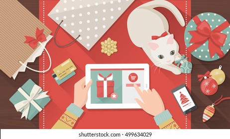 Woman purchasing Christmas gifts online using a tablet, her cat is playing with a bauble on the desk, holiday and celebrations banner