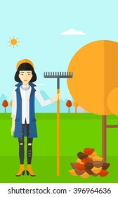 Woman with rake standing near tree and heap of autumn leaves.
