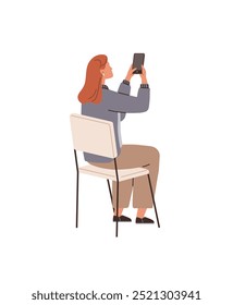 A woman with red hair sits on a chair, holding a smartphone to take a photo. She is casually dressed in a blue jacket and brown pants. Ideal for technology, social media, communication, lifestyle