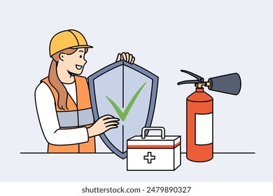 Woman safety specialist, stands near table with first aid kit and fire extinguisher and holds shield in hands. Girl safety inspector calls for caution, working to preserve life and health.