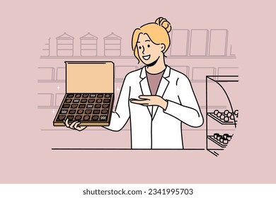 Woman seller with box of chocolates works in confectionery store and offers to buy appetizing dessert made from milk and cocoa. Girl in white coat of seller demonstrates chocolates with smile